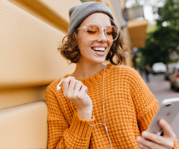 a smiling woman in a gray cap, glasses and a yellow sweater holds a smartphone in her hand